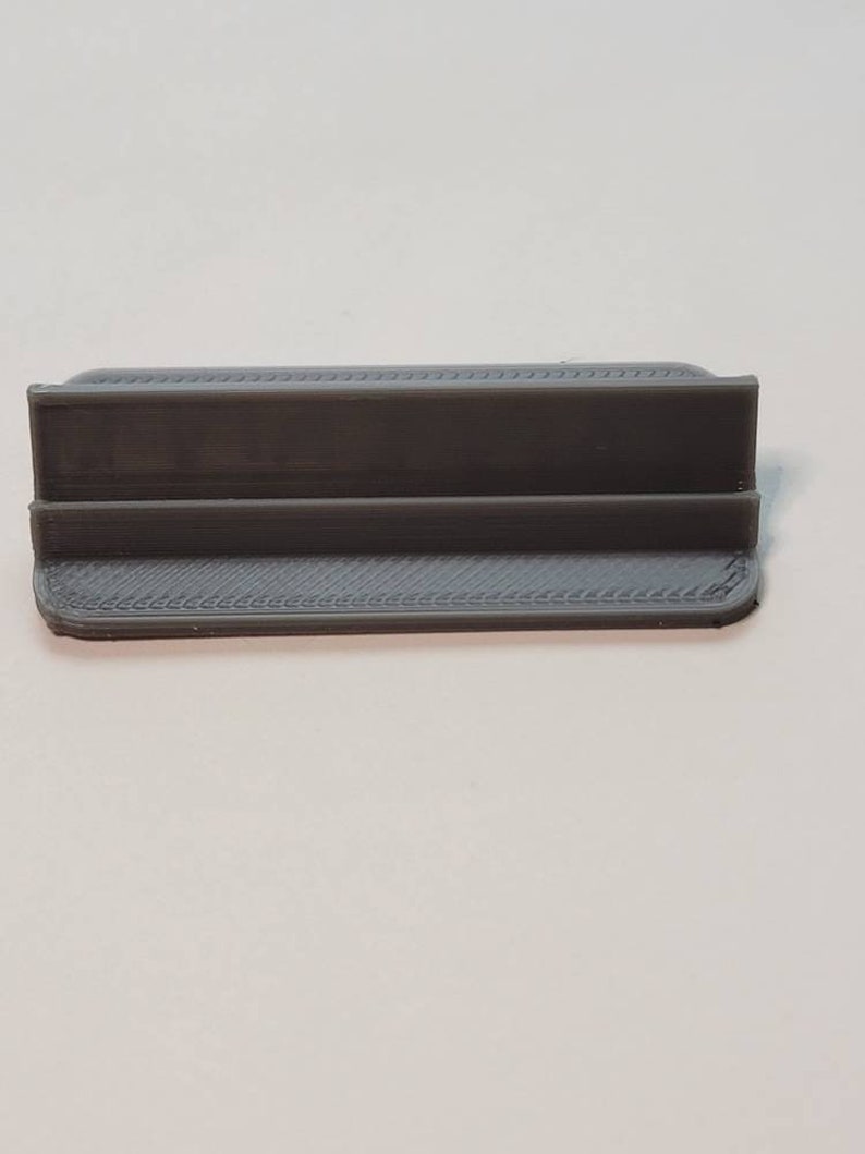 Pu&ograve; includere: Un supporto in plastica grigia stampato in 3D con una base rettangolare e una piattaforma rialzata al centro.
