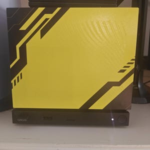 May include: A rectangular electronic device featuring a bright yellow front panel with black geometric angular patterns. The base is a black horizontal bar containing a small power button with a white indicator light, a USB port, and a small logo.