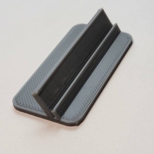 Pu&ograve; includere: Un supporto per telefono in plastica grigia con un design semplice. Il supporto ha due supporti verticali che creano uno spazio per appoggiare un telefono.