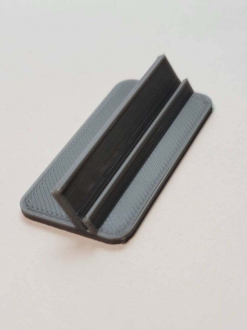 Pu&ograve; includere: Un supporto per telefono in plastica grigia con un design semplice. Il supporto ha due supporti verticali che creano uno spazio per appoggiare un telefono.