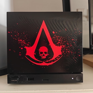 May include: A black rectangular electronic device featuring a prominent red graphic of a stylized crest with a skull in the center. The front panel includes a USB port, a small white indicator light, and a circular power button on the right side.