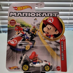 Pu&ograve; includere: Un'auto giocattolo Hot Wheels Mario Kart in metallo pressofuso con Baby Mario su un kart bianco e grigio con ruote gialle. La confezione presenta una bandiera a scacchi rossa e bianca, le parole "Baby Mario" e "B-Dasher", e un'immagine di Baby Mario con un berretto rosso e una salopette.