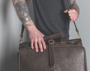 May include: A brown leather messenger bag with a long adjustable strap. The bag has a buckle closure and a flap with a metal clasp. The bag is worn by a person with a tattoo on their arm.