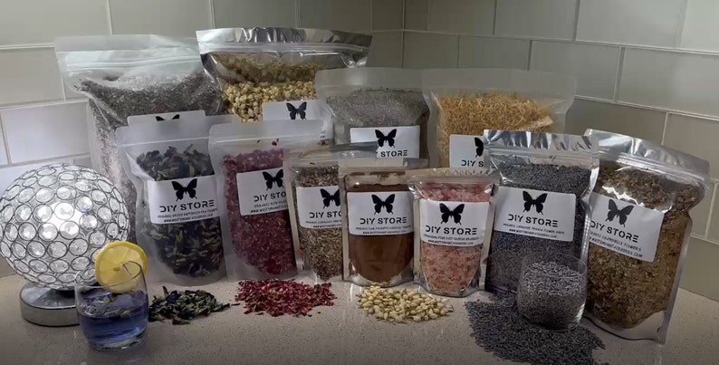 May include: A variety of dried flowers and herbs in clear plastic bags with a butterfly logo and the text "DIY STORE". The bags are arranged on a countertop with a glass of blue liquid and a lemon wedge.