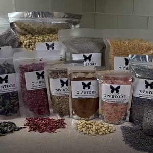 May include: Assortment of dried flowers and herbs in clear plastic bags, labeled "DIY STORE". Various colors and textures are visible, including lavender, rose petals, and other botanicals. A cocktail with a lemon slice sits in front of the bags.