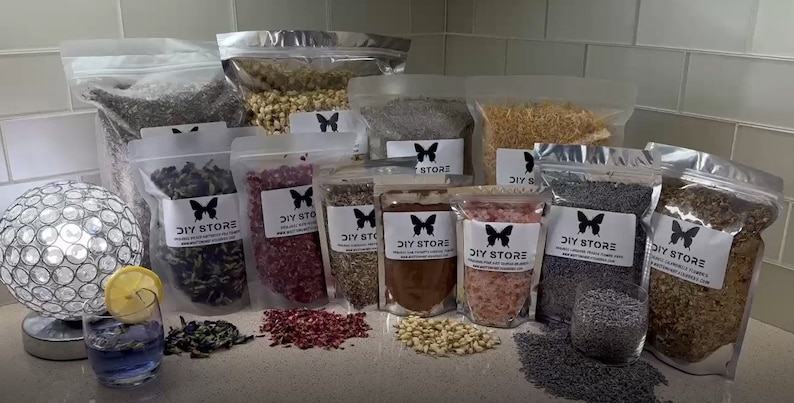 May include: Assortment of dried flowers and herbs in clear plastic bags, labeled "DIY STORE". Various colors and textures are visible, including lavender, rose petals, and other botanicals. A cocktail with a lemon slice sits in front of the bags.