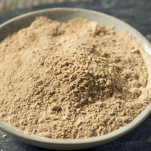 Peut inclure: Une poudre brun clair remplit un bol en céramique grise, avec un petit tas de la même poudre visible à gauche. La poudre a une texture fine et granuleuse. Le bol repose sur une surface sombre et texturée.