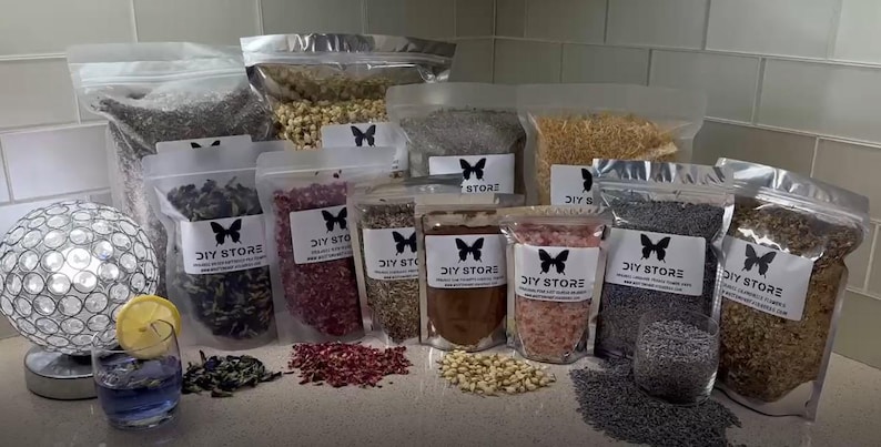 May include: Assortment of dried herbs and botanicals in clear, resealable bags, labeled "DIY STORE" with a butterfly logo. Various colors and textures are visible, including lavender, rose petals, and other dried ingredients. A cocktail with a lemon slice is in the foreground.
