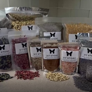 May include: Assortment of dried flowers and herbs in clear plastic bags, labeled "DIY STORE". Various colors and textures are visible, including lavender, rose petals, and other botanicals. A cocktail with a lemon slice sits in front of the bags.