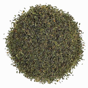 May include: A circular pile of dried, finely chopped green mint leaves. The texture is flaky and varied in shades of dark and forest green, with small brown flecks throughout. The herbs are arranged in a dense, uniform mound against a white background.