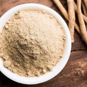 May include: A white ceramic bowl filled with a mound of fine, light beige ashwagandha powder. Beside the bowl lie several dried, woody, light brown ashwagandha roots. The texture of the powder is granular and uniform, contrasting with the fibrous roots.