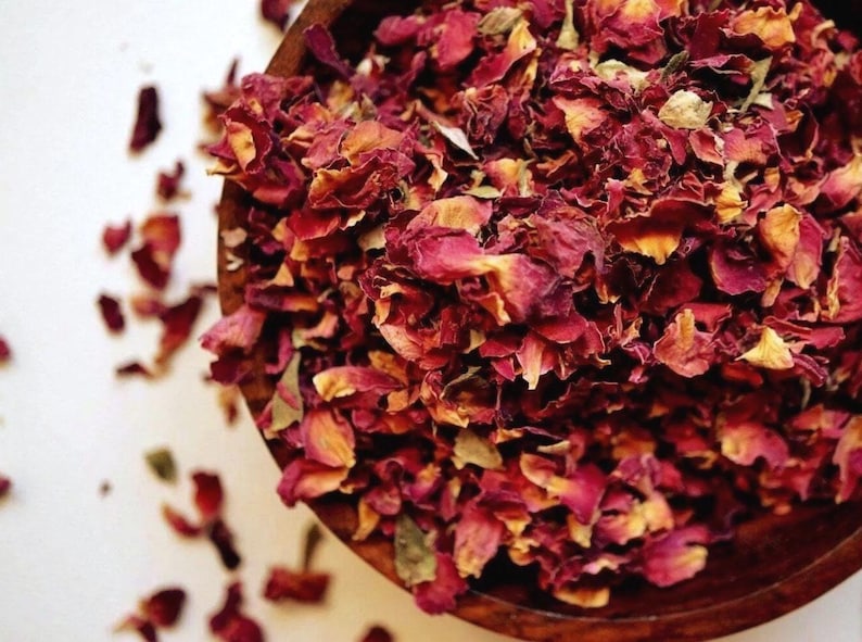 May include: A wooden bowl filled with dried rose petals. The petals are a mix of deep red and faded yellow, with some scattered outside the bowl. The image is taken from a high angle, showing the texture and color of the petals.