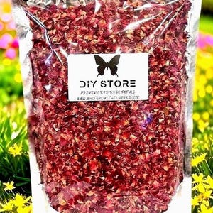 May include: A clear plastic bag filled with dried red rose petals. A white label with a black butterfly logo reads "DIY STORE PREMIUM RED ROSE PETALS WWW.WHITEMOUNTAINHERBS.COM". The background is a blurred view of yellow and green foliage.