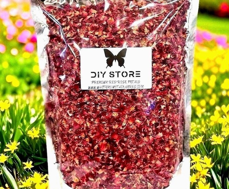 May include: A clear plastic bag filled with dried red rose petals. A white label with a black butterfly logo reads "DIY STORE PREMIUM RED ROSE PETALS WWW.WHITEMOUNTAINHERBS.COM". The background is a blurred view of yellow and green foliage.