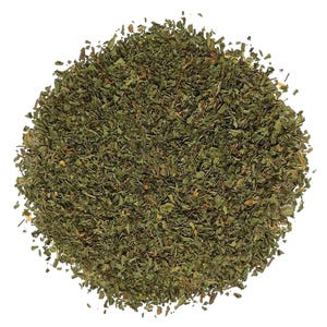May include: A circular pile of dried, finely chopped green herbs. The texture is flaky and varied, featuring shades of dark forest green, olive, and occasional flecks of light brown. The herbs are arranged in a dense, uniform mound against a white background.