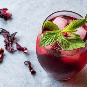 May include: A glass of red hibiscus tea with ice and fresh mint leaves. The tea is a deep red color and the mint leaves are green.
