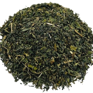 May include: A pile of dried, green herbs against a white background. The herbs appear to be finely chopped and have a slightly coarse texture. The color is a deep, natural green, with some lighter and darker variations throughout the pile. The image is well-lit, highlighting the details of the herbs.