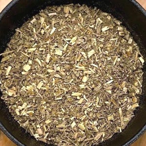 May include: Close-up of dried herbs in a black bowl. The herbs are a mix of green, tan, and brown, with a variety of textures. The bowl is round and has a slightly rough texture. The image is well-lit, showing the details of the herbs.