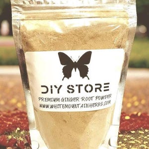 May include: A clear plastic bag filled with light brown powder. A white label features a black butterfly and the words "DIY STORE" above "PREMIUM GINGER ROOT POWDER" and a website address. The background is blurred with green and red tones.