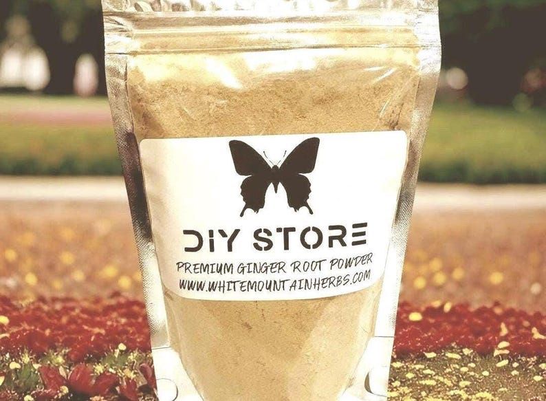 May include: A clear plastic bag filled with light brown powder. A white label features a black butterfly and the words "DIY STORE" above "PREMIUM GINGER ROOT POWDER" and a website address. The background is blurred with green and red tones.