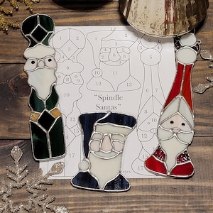 May include: A stained glass pattern for making three "Spindle Santas" ornaments. The pattern includes numbered pieces for each Santa, with a green, blue, and red color scheme.