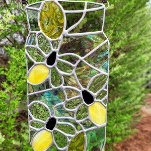 May include: Stained glass jar-shaped suncatcher with a brown hanging cord. The jar features yellow and black flower designs, with green and iridescent glass panels. The suncatcher is designed to hang and catch the light.