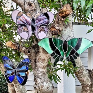 May include: Three stained glass butterfly ornaments hanging from a tree branch. The butterflies are purple, green, and blue.