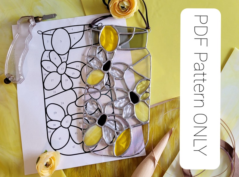 May include: A stained glass floral design with yellow and black flower details. The design is in a jar shape, with a PDF pattern and glass cutting tools nearby. The flowers have yellow petals and black centers. The glass is outlined with silver solder.
