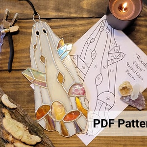 May include: A stained glass ghost with colourful accents and a black hanging cord. The ghost design features a pattern of mushrooms and geometric shapes. A pattern for the stained glass ghost is also visible, along with a lit candle.
