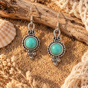 Può includere: Un paio di orecchini pendenti con pietre rotonde color turchese incastonate in metallo color argento anticato. Ogni orecchino presenta una cornice circolare con un bordo decorativo smerlato, un occhiello in alto e tre piccole sfere argentate.