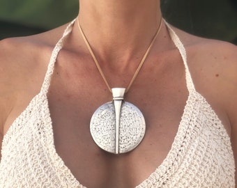 Puede incluir: Un collar con un colgante de plata, redondo y texturizado. El colgante tiene un pequeño diseño vertical puntiagudo en el centro. El collar lo lleva una persona con un tono de piel marrón claro. La persona lleva un top halter de crochet blanco.