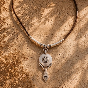 May include: A necklace featuring a dark brown leather cord with silver-toned metal beads and a central pendant. The pendant consists of a circular disc with a small flower motif, connected to a dangling, diamond-shaped charm with leaf engravings.