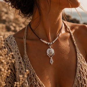 May include: A necklace featuring a dark cord and silver-toned metal components. The pendant consists of a circular disc with a central flower motif, connected to a dangling, diamond-shaped charm with an engraved pattern and a small dark stone.