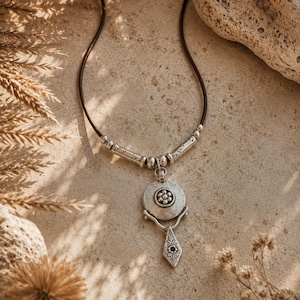 May include: A necklace featuring a dark brown cord with silver-toned metal beads and a circular pendant. The pendant displays a central flower motif and is attached to a dangling, diamond-shaped charm with engraved floral details and a dark center.