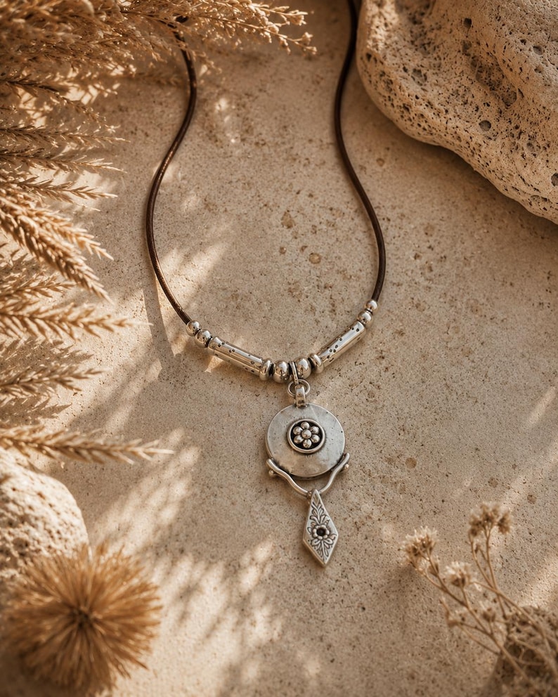 May include: A necklace featuring a dark brown cord with silver-toned metal beads and a circular pendant. The pendant displays a central flower motif and is attached to a dangling, diamond-shaped charm with engraved floral details and a dark center.