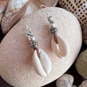 Può includere: Orecchini a conchiglia con dettagli in metallo color argento. Ogni orecchino presenta una conchiglia bianca pendente da un gancio argentato, con una perlina decorativa argentata e un cordino in pelle marrone. Gli orecchini sono esposti su una pietra chiara.