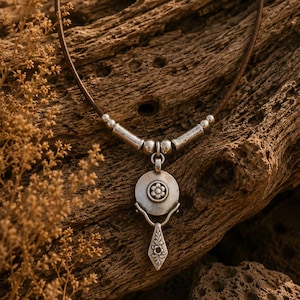 May include: A necklace featuring a dark brown leather cord with silver-toned metal accents and a circular pendant. The pendant displays a small embossed flower at its center, with a dangling, diamond-shaped charm featuring intricate leaf engravings.