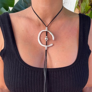 Puede incluir: Un collar con un colgante de plata con un diseño circular abstracto. El collar está hecho de cordón de cuero y tiene un acento de cuentas de plata.