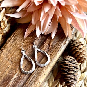 Può includere: Un paio di orecchini pendenti in argento con una forma a goccia attorcigliata. Gli orecchini sono su una superficie di legno con un fiore rosa e pigne.