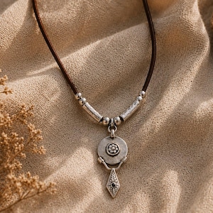 May include: A necklace featuring a dark brown leather cord with silver-toned metal accents. The pendant consists of a circular disc with a central flower motif, connected to a dangling, diamond-shaped charm with a small dark inlay at its center.