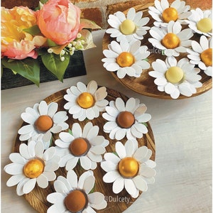 May include: A wooden tray with white daisy-shaped holders filled with chocolate eggs. Some eggs are covered in gold foil, while others are white. The tray is on a wooden table with a brick wall in the background.