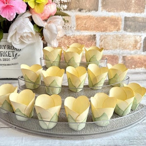 May include: A set of 18 yellow tulip-shaped paper cupcake liners with green leaf accents. The liners are arranged in a tiered display on a clear plastic stand.