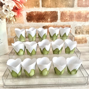 May include: Three tiers of clear acrylic cupcake stands, each holding white cupcake liners with green leaf designs. The liners are arranged in a staggered pattern.