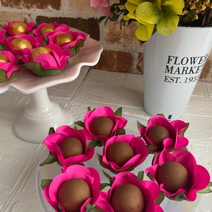 May include: Chocolate truffles wrapped in pink paper roses with green leaves. The truffles are arranged on a white plate with a white pedestal. A white vase with the text "FLOWER MARKET EST. 195" is in the background.