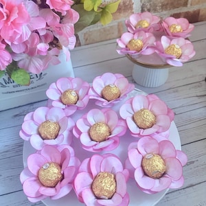 May include: A white plate holds several pink paper flower candies, each with a gold-wrapped chocolate center. Additional flower candies are on a small pedestal. Pink flowers are in the background.