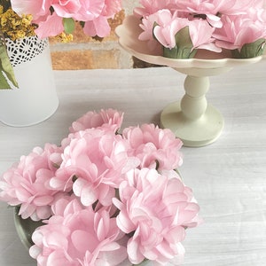 May include: A group of pink paper flowers arranged on a white pedestal cake stand. The flowers are made of delicate, layered paper and have a soft, romantic look.