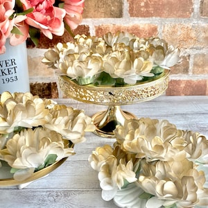 May include: Three gold-rimmed plates with white paper flowers on a whitewashed wood surface. The flowers are arranged in a circular pattern and have a subtle gold sheen. The plates are on a whitewashed wood surface.