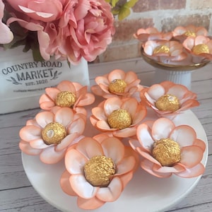 May include: A white plate holds several peach-colored paper flowers, each with a gold-wrapped chocolate candy in the center. A bouquet of pink flowers sits in a white container with the text "COUNTRY ROAD MARKET EST. 1926".