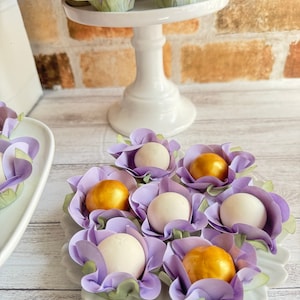 May include: Purple and green paper flower cups filled with white and gold chocolate candies. The candies are arranged on a white plate with a scalloped edge.