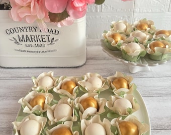 May include: A light green rectangular plate holds a dozen white and gold candies, each in a flower-shaped wrapper. A white ceramic container with pink flowers and the text "COUNTRY ROAD MARKET EST. 1926" is in the background.